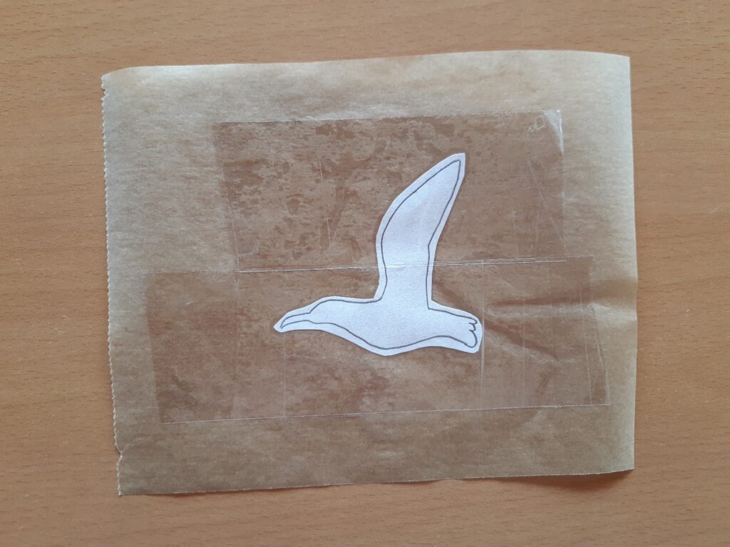 Dessin du contour d'un oiseau entre 2 bouts de scotchs sur une feuille de papier cuisson. Le tout posé sur une table en bois.